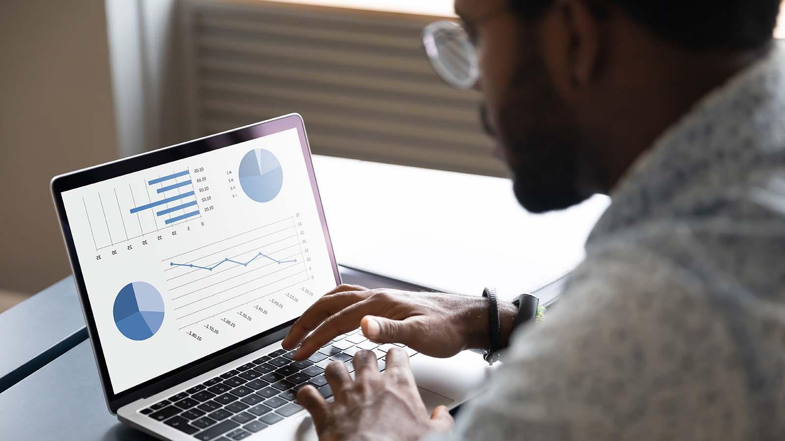 Focused young african american male professional working on project report by analyzing graphs and charts, sitting at table in office or home.